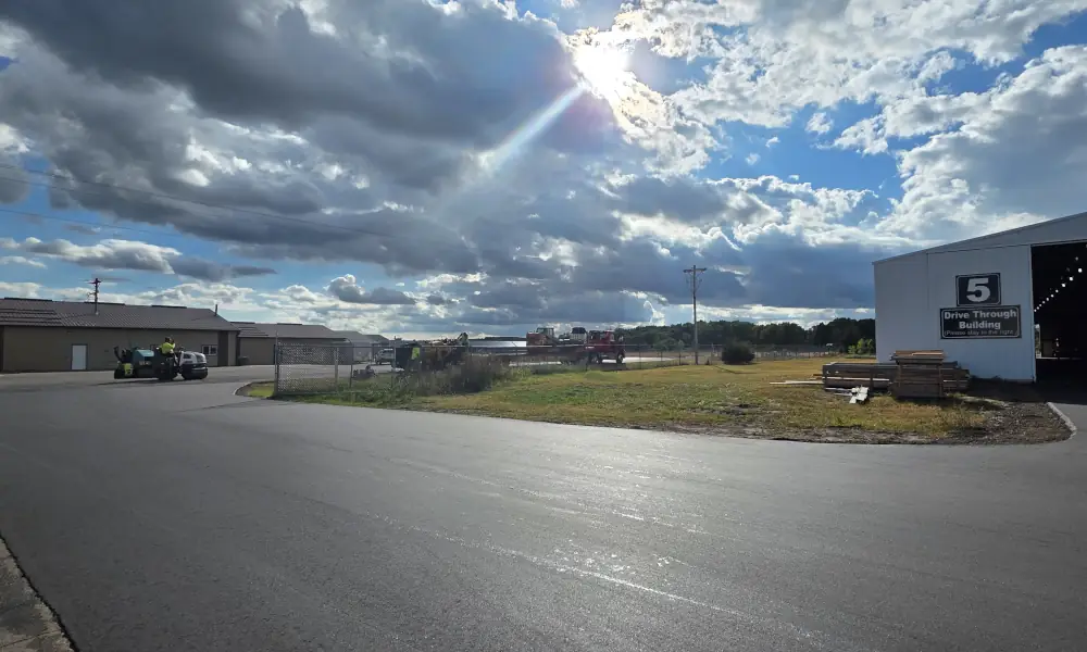 Newly paved asphalt parking lot at Marv’s True Value in Princeton, Minnesota, with construction equipment in the distance and sunlight breaking through the clouds.