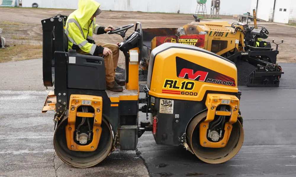 Worker operating a Mauldin RL10 roller to compact freshly laid asphalt behind an Erickson Asphalt paving machine on a commercial lot.